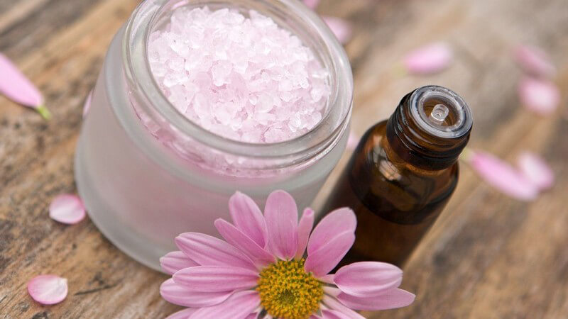 Nahaufnahme Döschen mit Badesalz, daneben kleine Flasche Duftöl und rosane Blüte