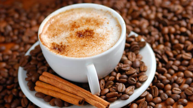 Cappuccinotasse mit Zimtschaum umgeben von Kaffeebohnen und Zimstangen