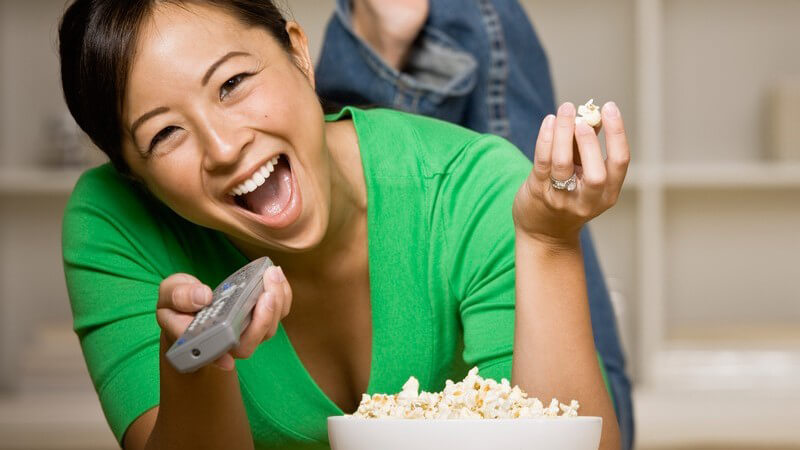 Junge, lachende Frau liegt mit Schüssel Popcorn auf dem Bauch und schaut Fern