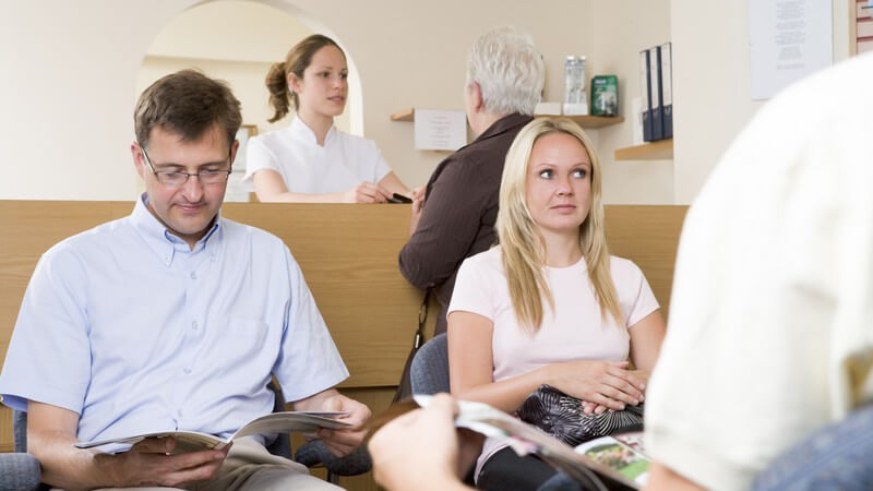 Patienten sitzen im Wartezimmer, im Hintergrund Arzthelferin an Anmeldung