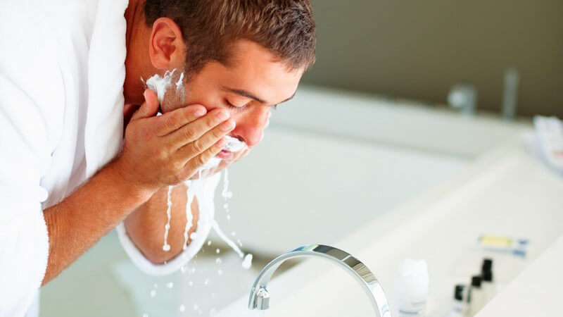 Mann wäscht sich am Waschbecken Rasierschaum aus Gesicht