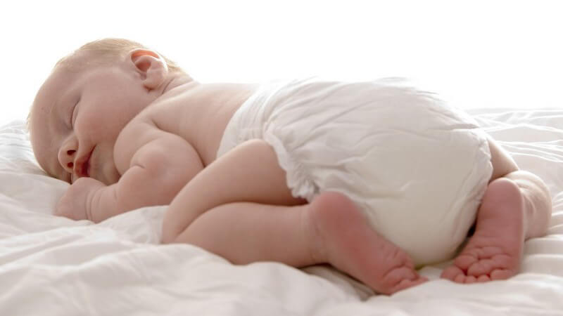 Säugling mit Windel liegt auf dem Bauch mit angezogenen Beinchen und schläft auf weißer Decke