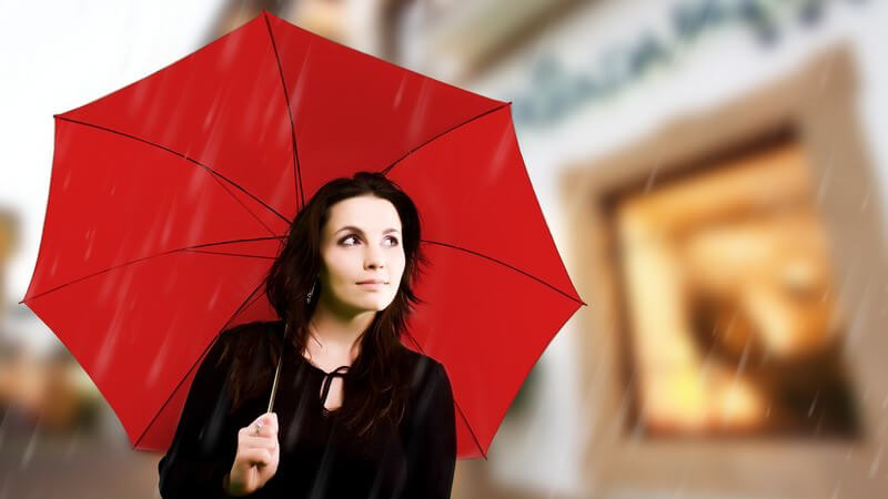 Dunkelhaarige Frau geht unter rotem Regenschirm an Regentag durch Stadt