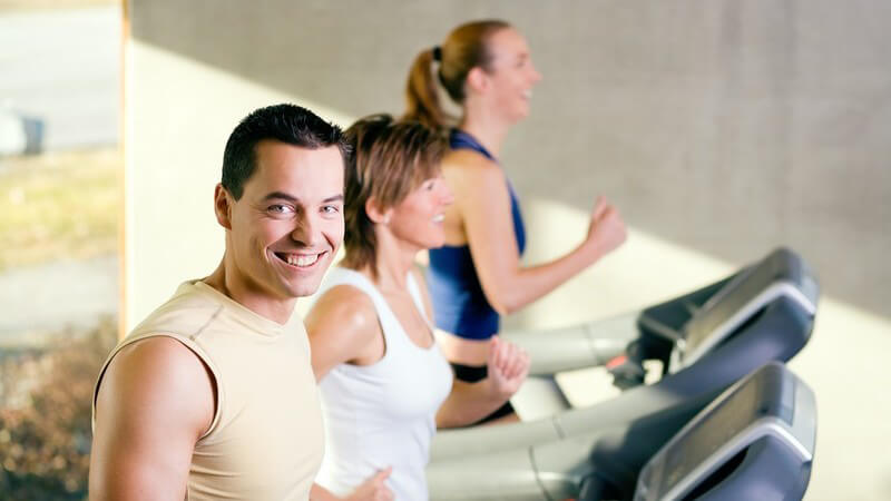 Mann auf Laufband im Fitnessstudio, im Hintergrund zwei Frauen auf Laufband
