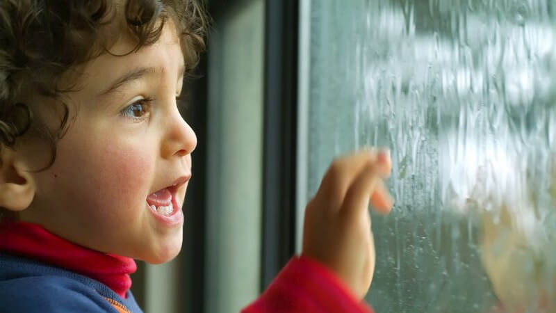 Kleiner Junge steht am Fenster und schaut dem Regen zu