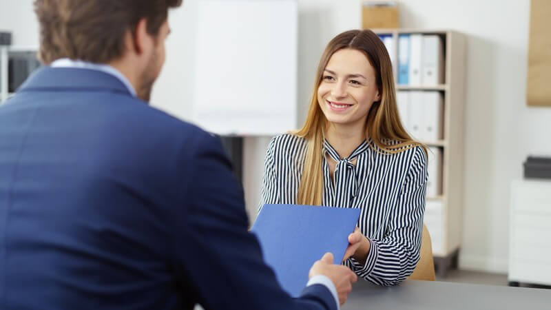 Junge Frau sitzt lächelnd einem jungen Arbeitgeber gegenüber, der die blaue Bewerbungsmappe entgegen nimmt