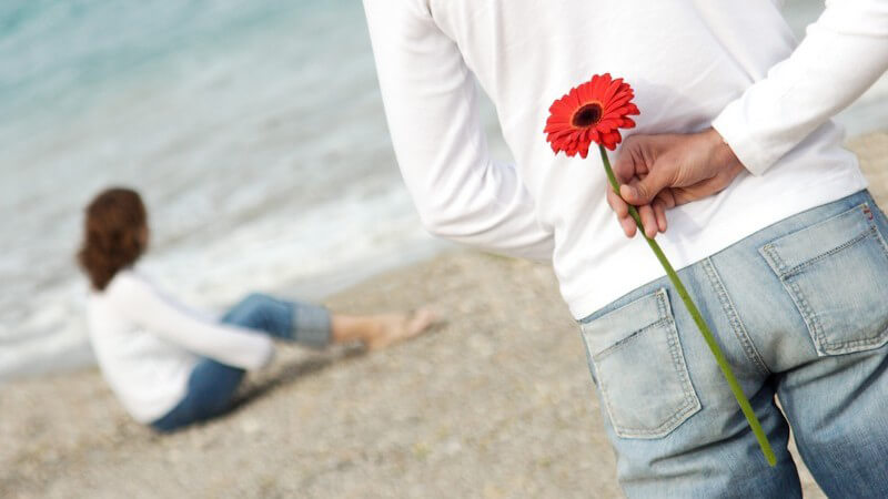 Junger Mann am Strand hält eine rote Blume hinter seinem Rücken versteckt