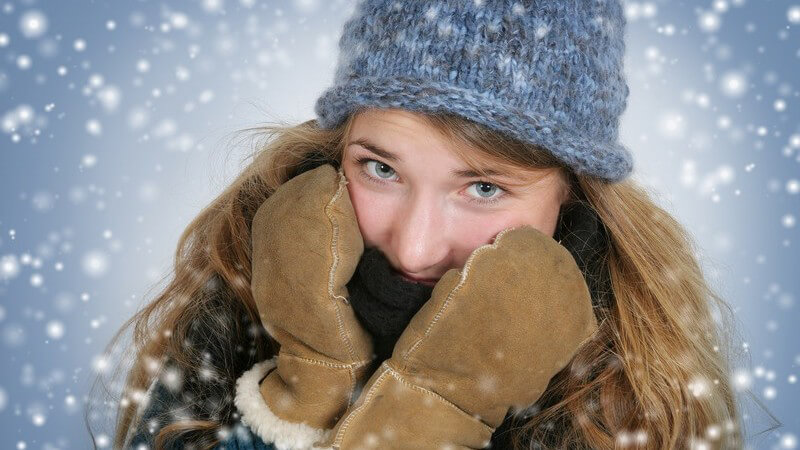 Es schneit- Junge Frau mit braunen Fäustlingen und hellblauer Mütze zieht sich schwarzen Schal über Mund