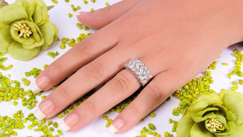 Hand mit langen, gepflegten Fingernägeln und funkelndem Ring liegt neben einer grünen Blüte