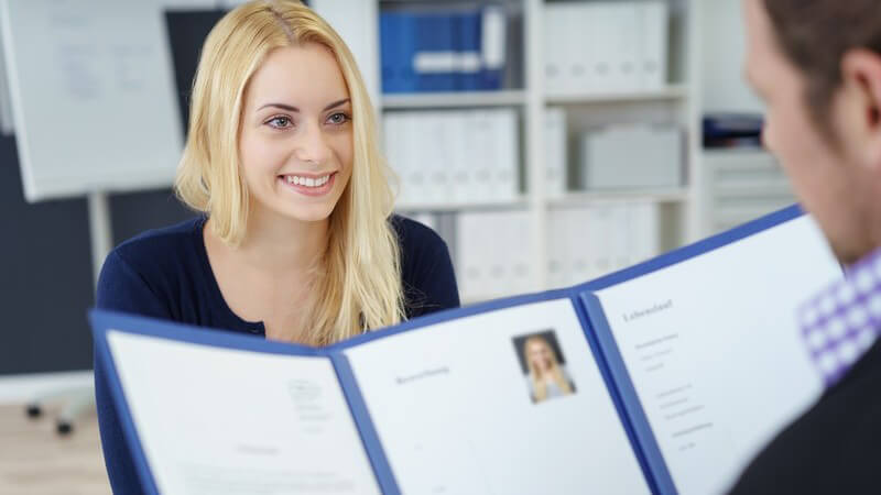 Junge blonde Frau sitzt lächelnd einem Arbeitgeber gegenüber, der sich die Bewerbungsmappe ansieht