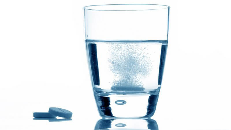 Links zwei Tabletten übereinander, rechts Wasserglas mit Wasser und sprudelnder Tablette, Kopfschmerztabletten