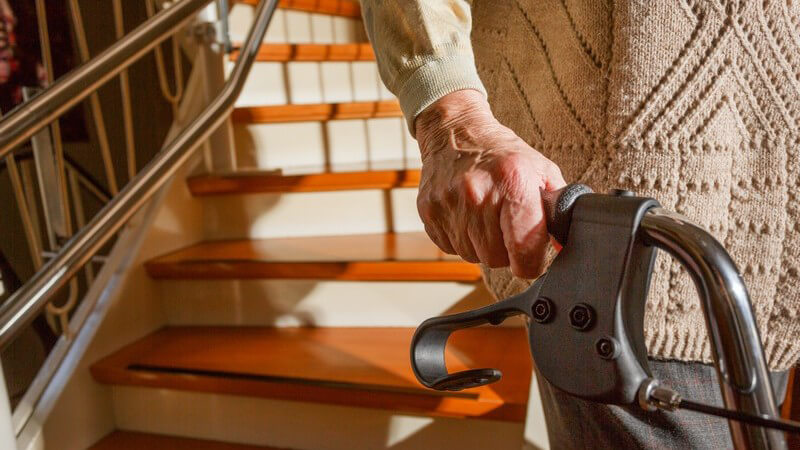 Hand einer alten Frau am Rollator, im Hintergrund Treppe ihrer Wohnung