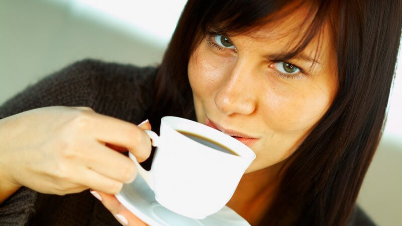 Junge brünette Frau schaut in Kamera und trinkt an Tasse Kaffee