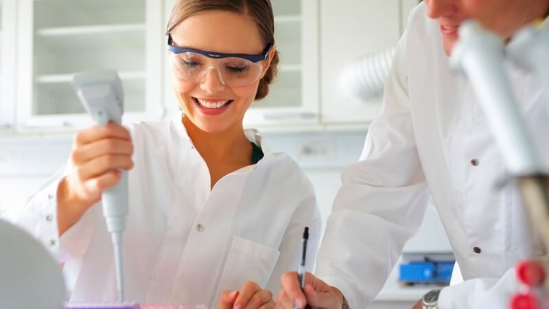 Wissenschaftlerin in weißem Kittel und Schutzbrille bei der Arbeit im Labor
