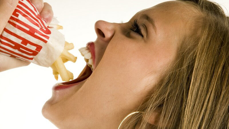Junge Frau hält Tüte Pommes über offenen Mund, weißer Hintergrund