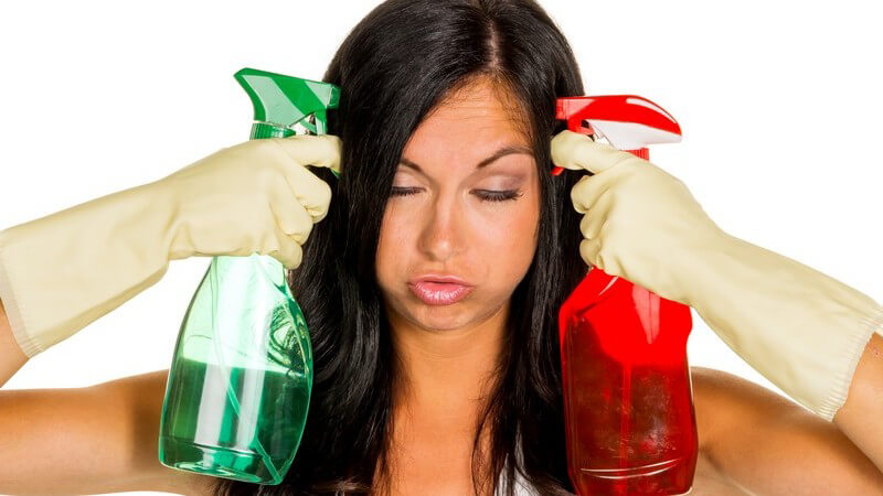 Frau im Putz-Stress hält sich zwei Sprühflaschen mit Reinigungsmittel an den Kopf