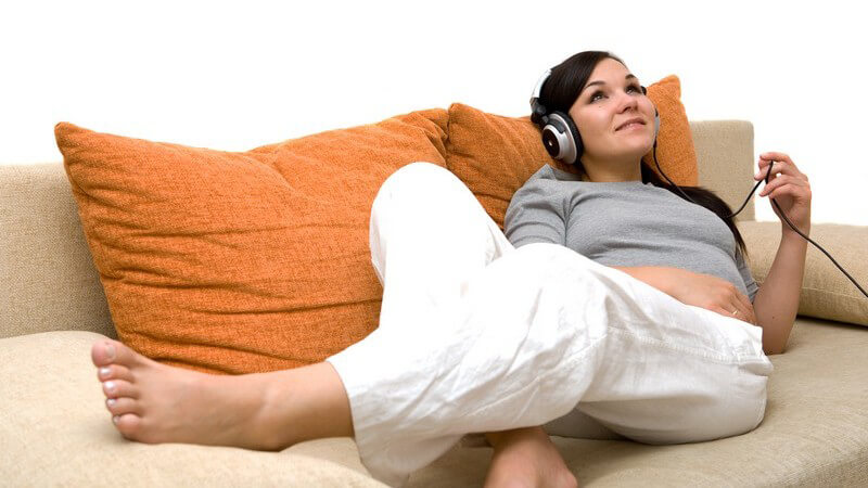 Junge Frau liegt barfuss auf Couch, hört Musik über Kopfhörer