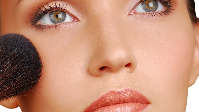 Frau mit hellem Lidschatten, grünen Augen und apricot Lippenstift hält Schminkpinsel an rechte Wange
