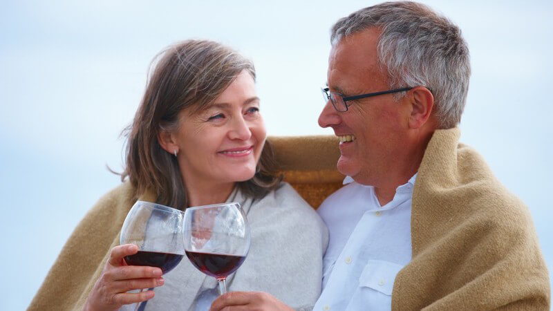 Älteres Paar sitzt in Decke gekuschelt und stößt mit Rotwein an