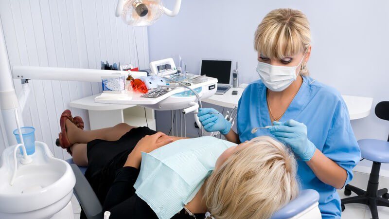 Patientin auf Liege im Behandlungsraum einer Zahnarztpraxis, daneben sitzt Zahnärztin mit Mundschutz