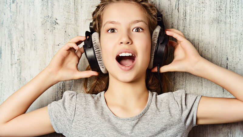 Mädchen in grauem Shirt hört Musik mit einem großen Kopfhörer und hat den Mund weit geöffnet