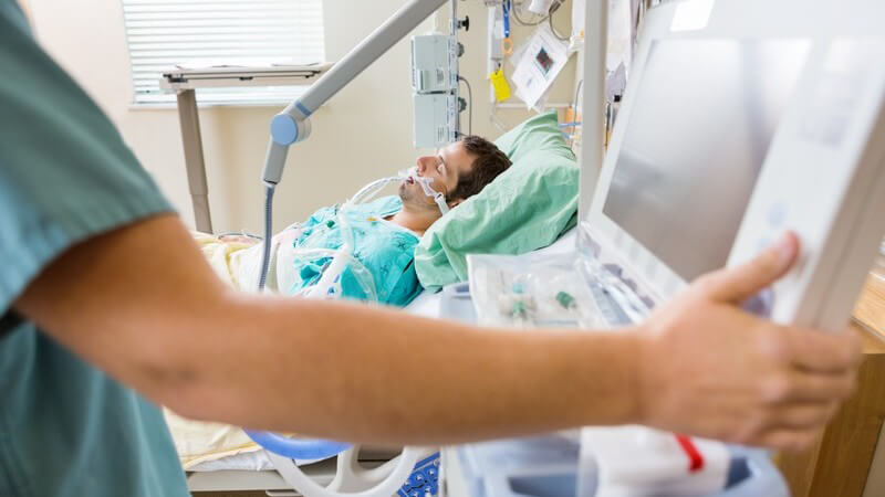 Krankenschwester/Gesundheits- und Krankenpflegerin bedient Monitor im Krankenhauszimmer, im Hintergrund Patient im Bett