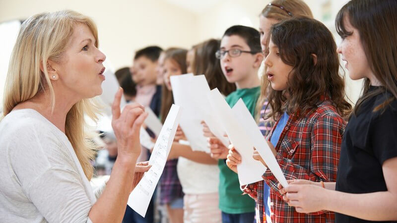 Kinder stehen nebeneinander, halten ein Notenblatt und singen im Chor, unterrichtet von einer blonden Lehrerin