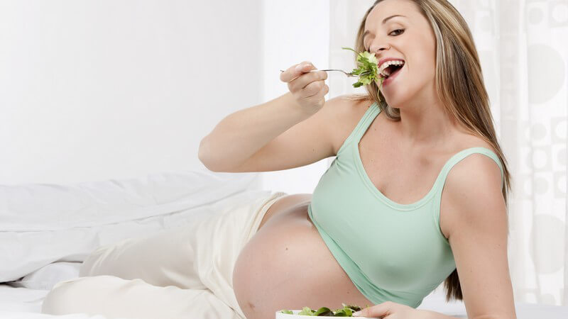 Schwangere Frau liegt auf Bett und isst Salat