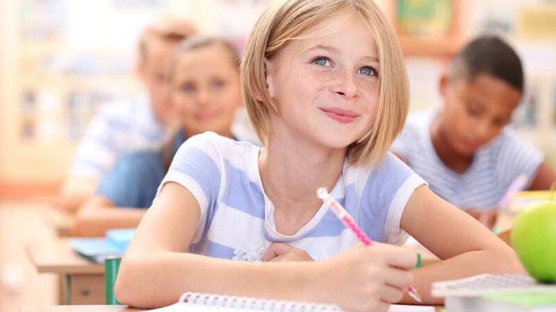Mädchen mit schulterlangen blonden Haaren und Sommersprossen sitzt mit Stift und Block am Tisch im Klassenzimmer