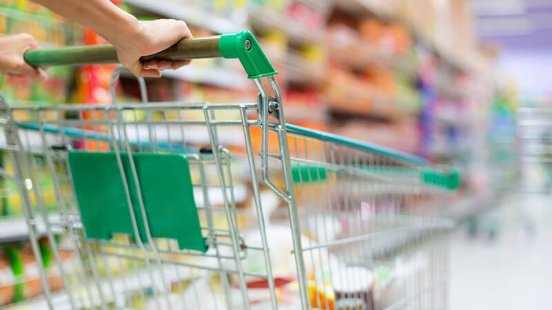 Einkaufswagen wird im Gang eines Supermarkts geschoben, im Hintergrund Regale mit Lebensmitteln