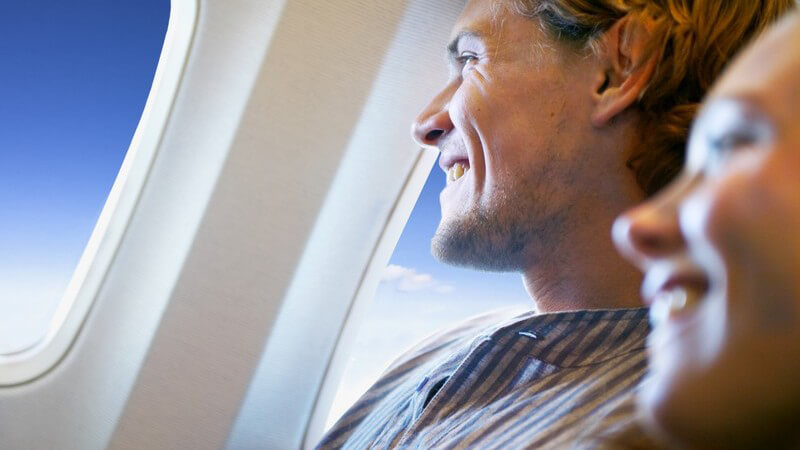 Junger Mann auf Fenstersitz in Flugzeug, schaut aus dem Fenster in den Himmel, junge lächelnde Frau unscharf daneben