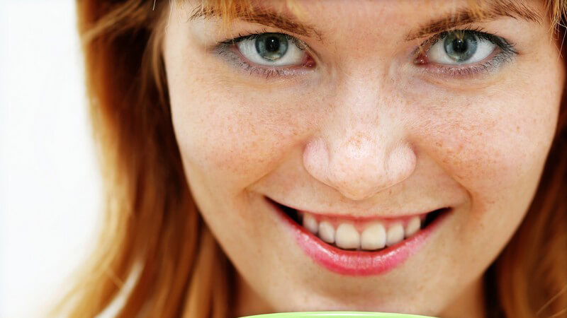 Rothaarige Frau mit Sommersprossen hält Kaffee und lächelt in die Kamera