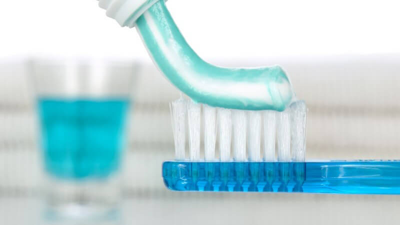 Zahncreme wird auf Zahnbürste aufgetragen, im Hintergrund Glas mit Mundspülung