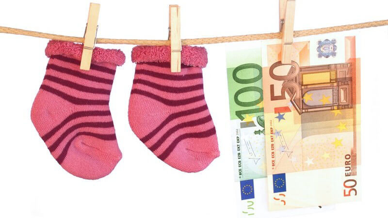Zwei pinke Babysöckchen und zwei Euro-Geldscheine hängen mit Holzwäscheklammern an einer Leine vor weißem Hintergrund