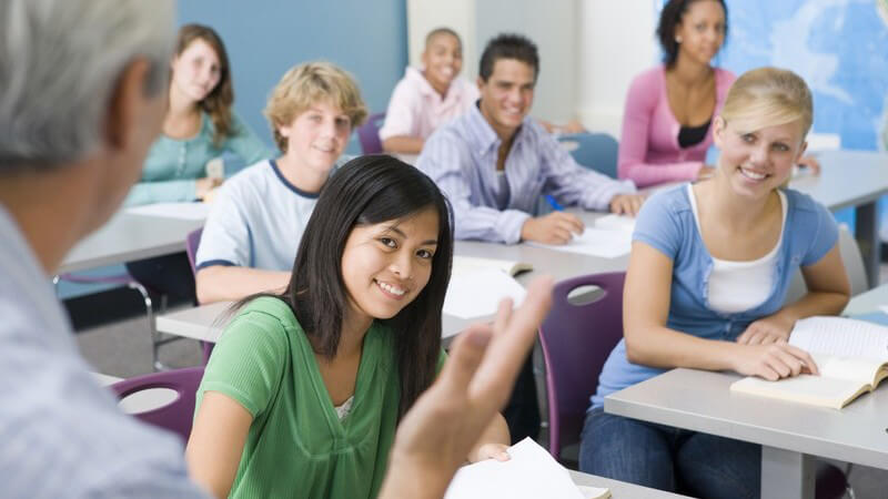 Lehrer steht vor seiner Schulklasse in Klassenraum, Schüler lächeln ihn an
