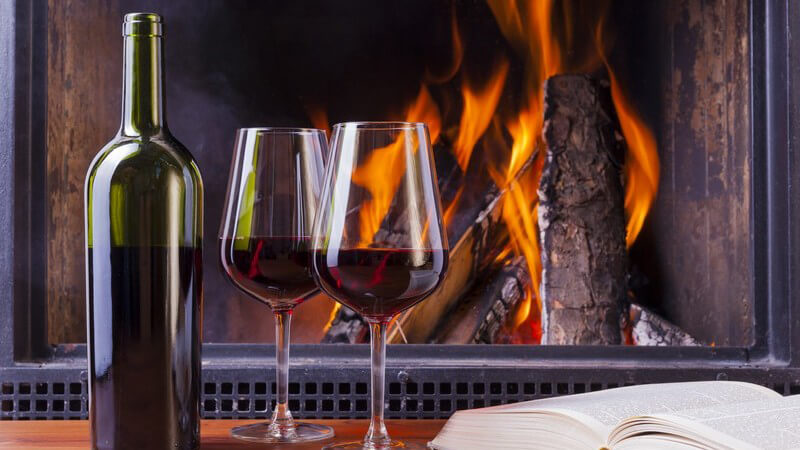 Blick auf Kaminfeuer, im Vordergrund Flasche und 2 Gläser Rotwein, aufgeschlagenes Buch