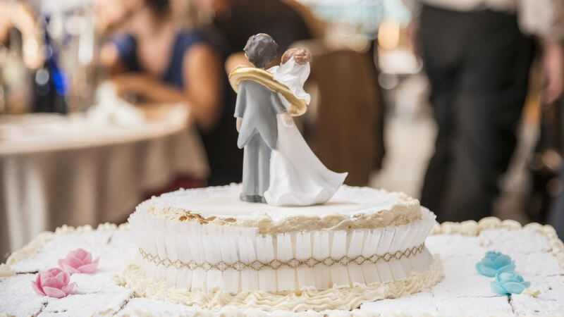 Hochzeitstorte mit Brautpaarfiguren als Topping, im Hintergrund sitzt die Hochzeitsgesellschaft