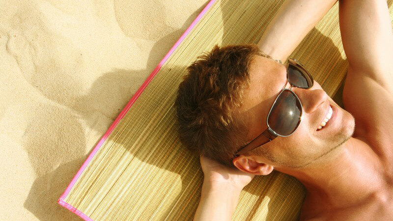Mann mit schwarzer Sonnenbrille verschränkt die Arme hinterm Kopf, liegt auf Strohmatte am Strand