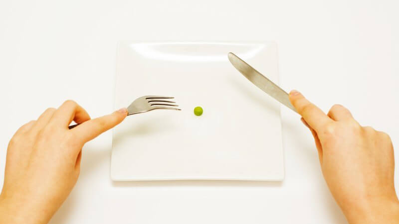 Diät - Zwei Hände mit Messer und Gabel an Teller, auf Teller liegt eine Erbse