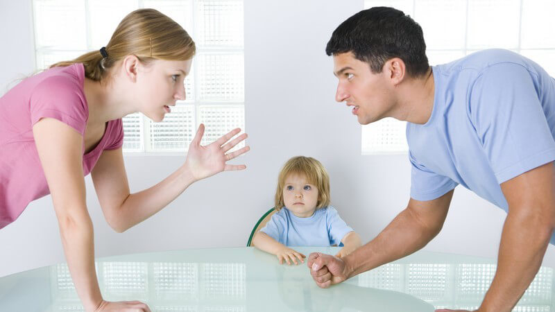 Junge Eltern auf Tischplatte gestützt streiten, dahinter sitzt kleines trauriges Mädchen am Tisch