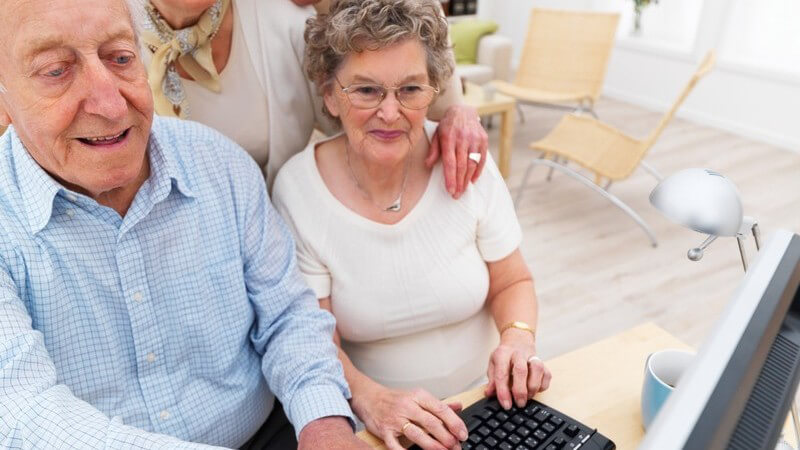 Gruppe alter Menschen sitzt/steht vor Computer, alle lachen