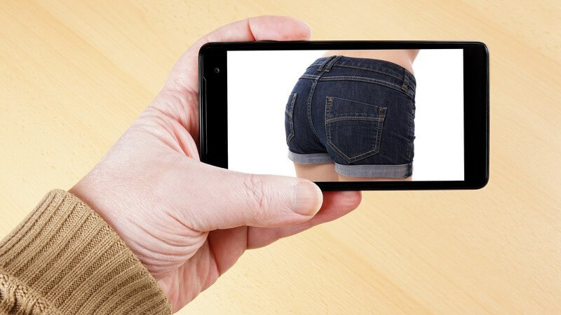 Mann hält ein Smartphone mit einem Foto eines Frauenhinterns in Hot Pants