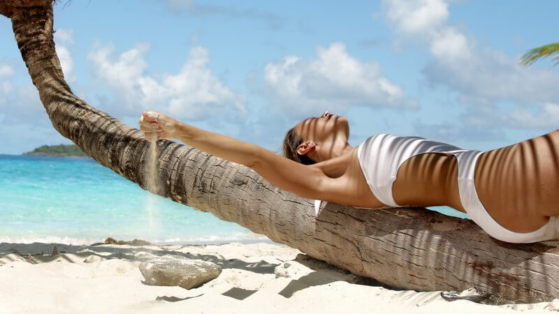 Hübsche Frau im Badeanzug liegt auf Palmenstamm und spielt mit Sand Monokini