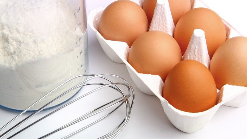 Sechs rohe Eier, daneben Glas mit Mehl und Schneebesen für Crêpes