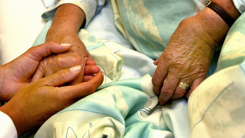 Pflegerin hält die faltige, rechte Hand einer bettlegerichen, alten Frau