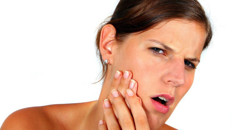 Junge Frau mit schmerzverzerrtem Gesicht hält sich beide Hände an Wange, Zahnschmerzen