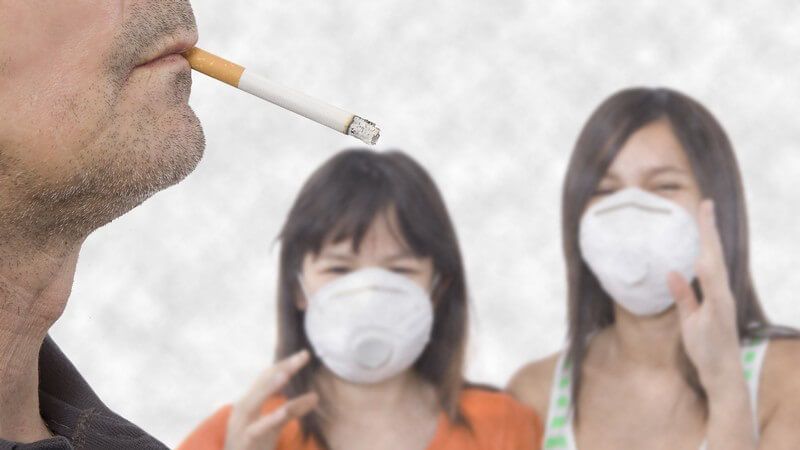 Zwei Mädchen mit Mundschutz vor Rauch und Qualm ärgern sich über rauchenden Mann mit Zigarette im Mund im Vordergrund