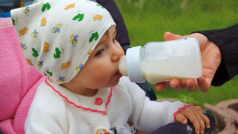 Kleinkind mit Mütze in Kinderwagen wird von Hand der Mutter mit einer Babyflasche gefüttert, im Hintergrund grüner Rasen