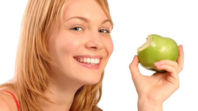 Blonde Frau lächelt und hält grünen, angebissenen Apfel neben ihr Gesicht