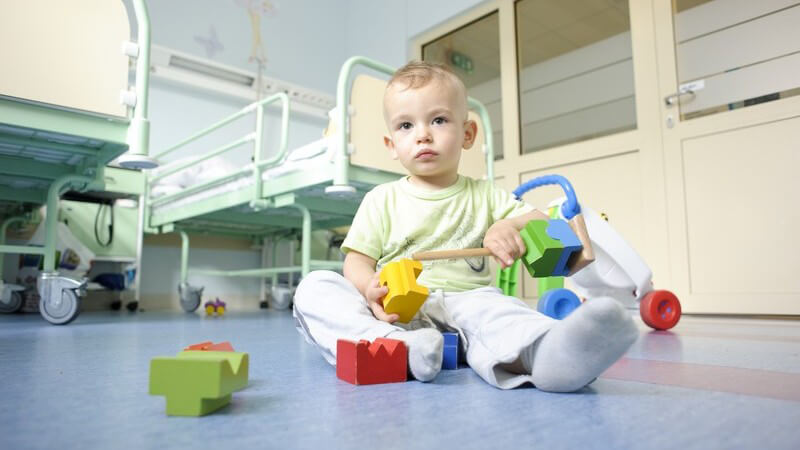 Kleiner Junge sitzt auf Boden im Krankenhaus und spielt mit Bauklötzen, im Hintergrund Krankenbetten